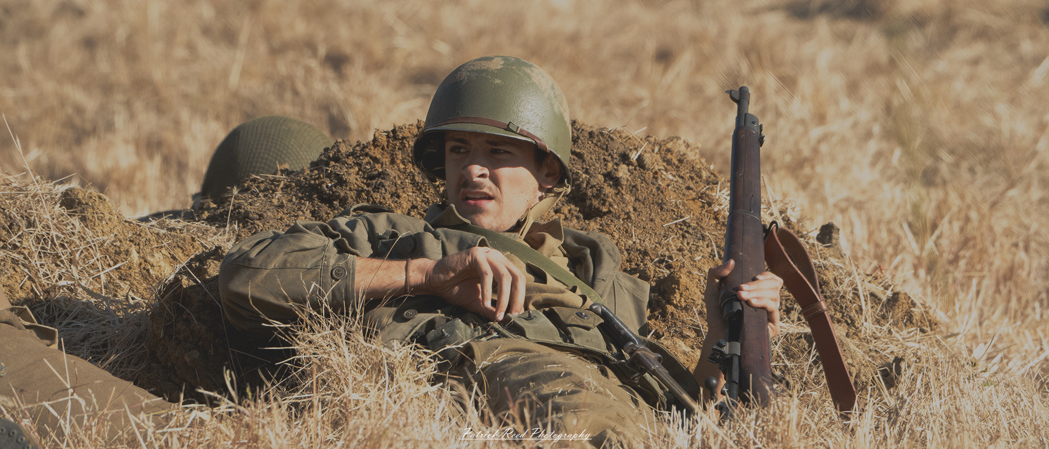 "Image of a soldier lying on his back in a field, reaching for more ammunition with one hand while gripping his rifle with the other. The soldier, dressed in military fatigues, has an intense expression as he prepares for the next engagement. The backdrop features a blend of tall grass and wildflowers under a clear blue sky, emphasizing the contrast between the serenity of nature and the soldier's urgent need for readiness."